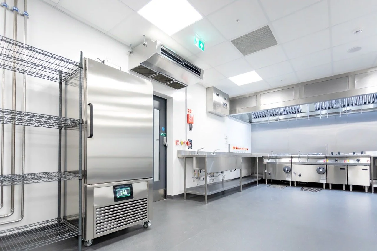 Glistening silver commercial kitchen with preparation tables and large silver fridges.