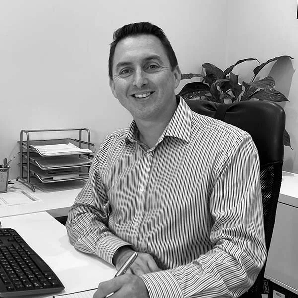 Black and white photo of a smiling man sitting at a desk in an office, with a potted plant and organised paper trays in the background.