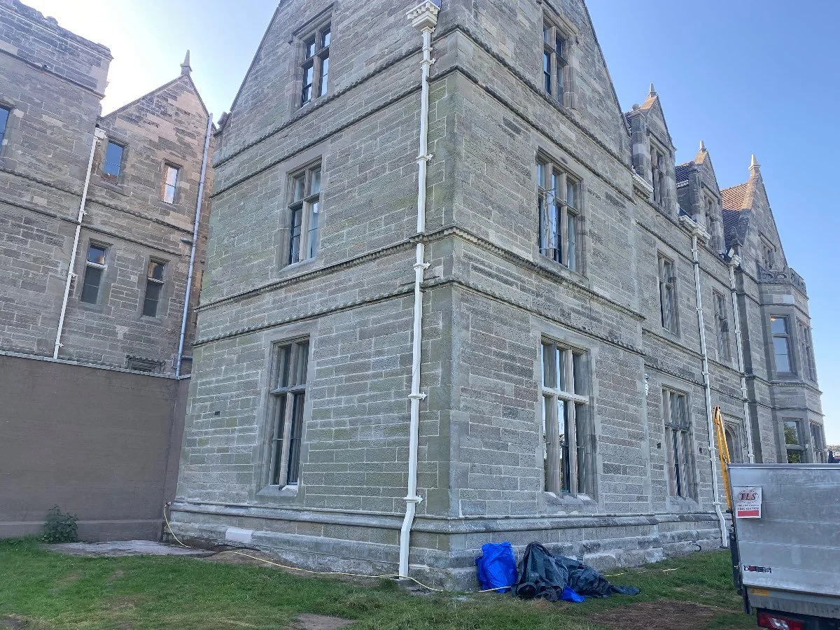 An old, large stone building with two stories on a sunny day. There is an area cornered-off by metal fencing suggesting building work is being completed.