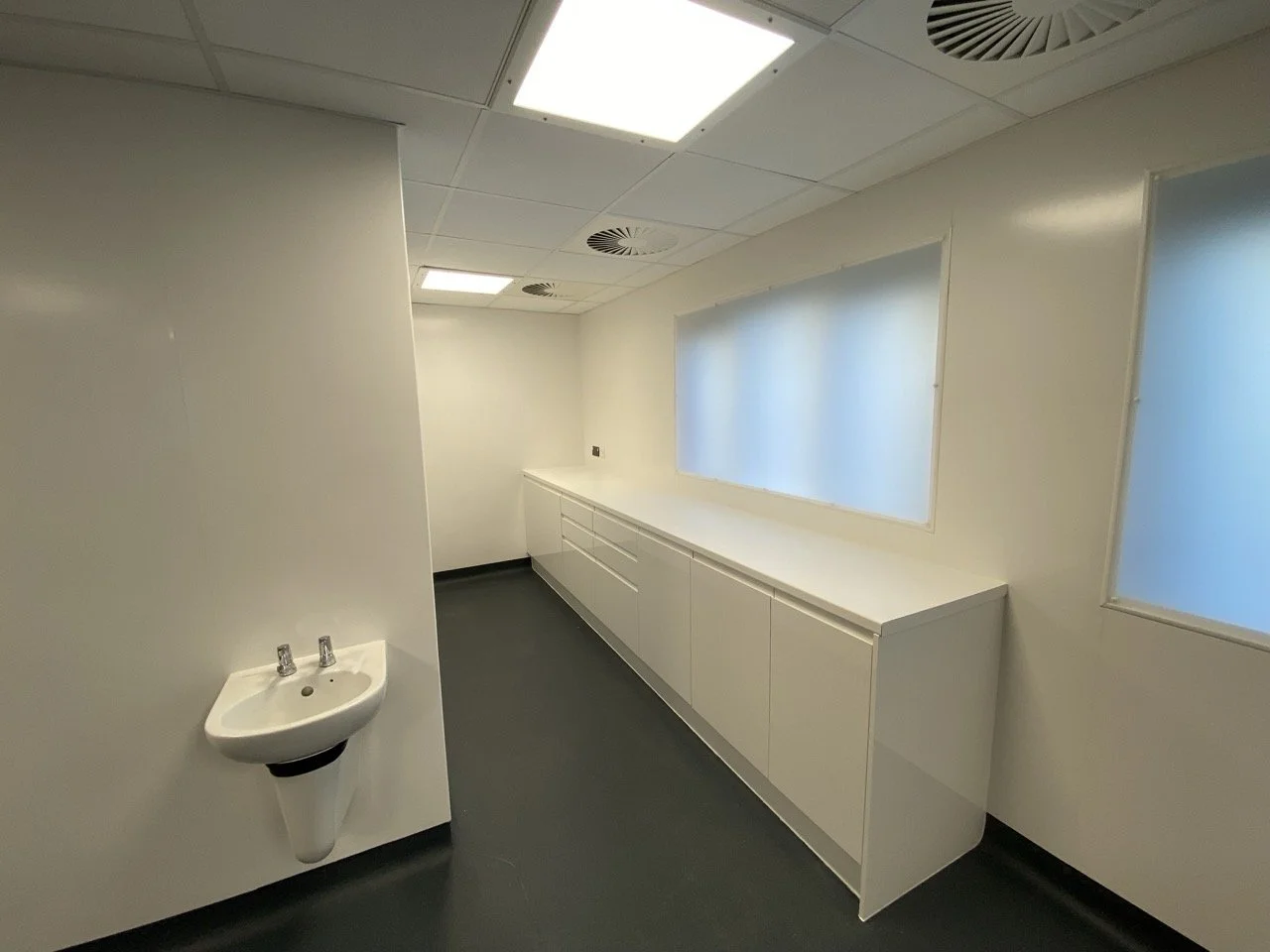 Clinical white toilet area in a health centre, there is a small white sink and row of white cupboards.