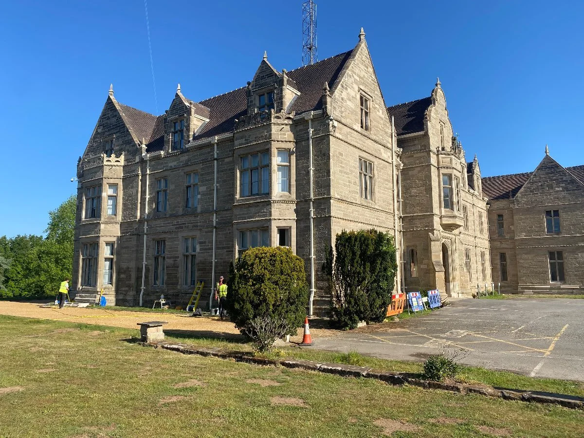 The side of a large two-story stone building with a large concrete driveway to the front and a grass lawn to the back.