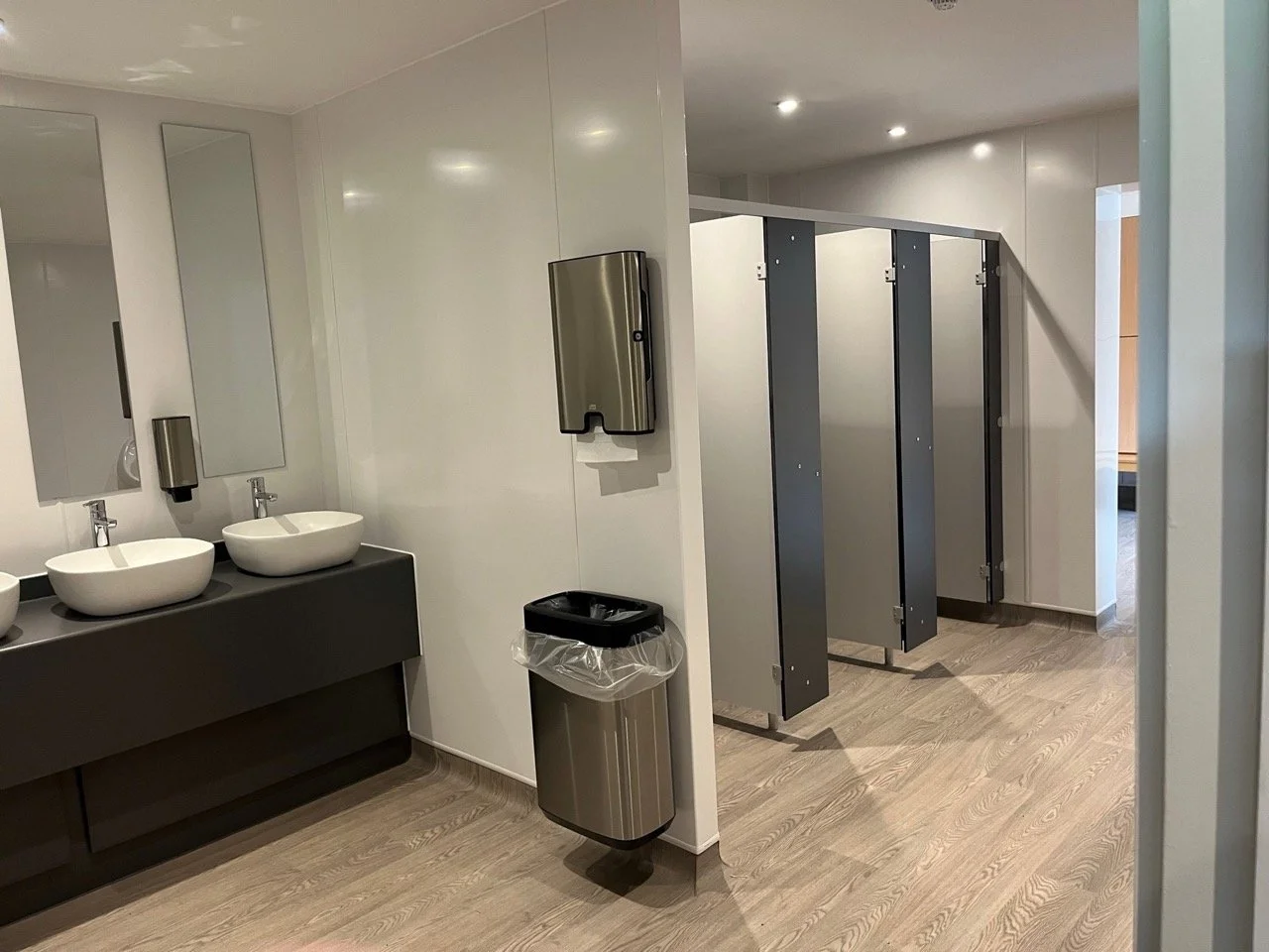 Hotel communal bathroom area with toilet stalls and two basins next to a bin and paper towel dispenser.