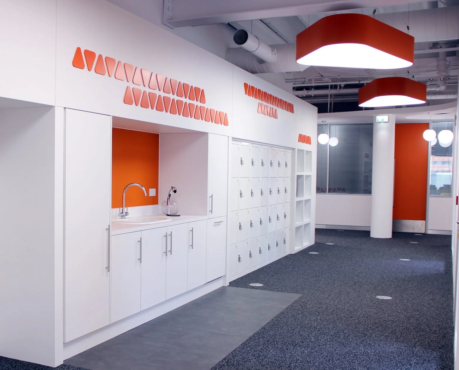 White-walled large room with white lockers and orange features including a splash back and large light shades.
