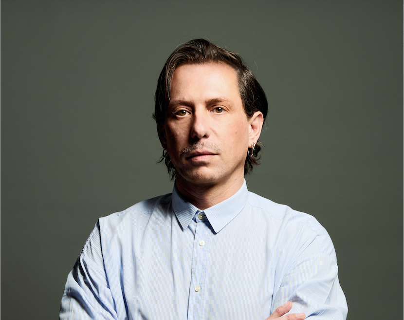 Un homme avec des cheveux longs et décolorés, portant une chemise à rayures, regarde sérieusement l'objectif avec les bras croisés, contre un fond gris