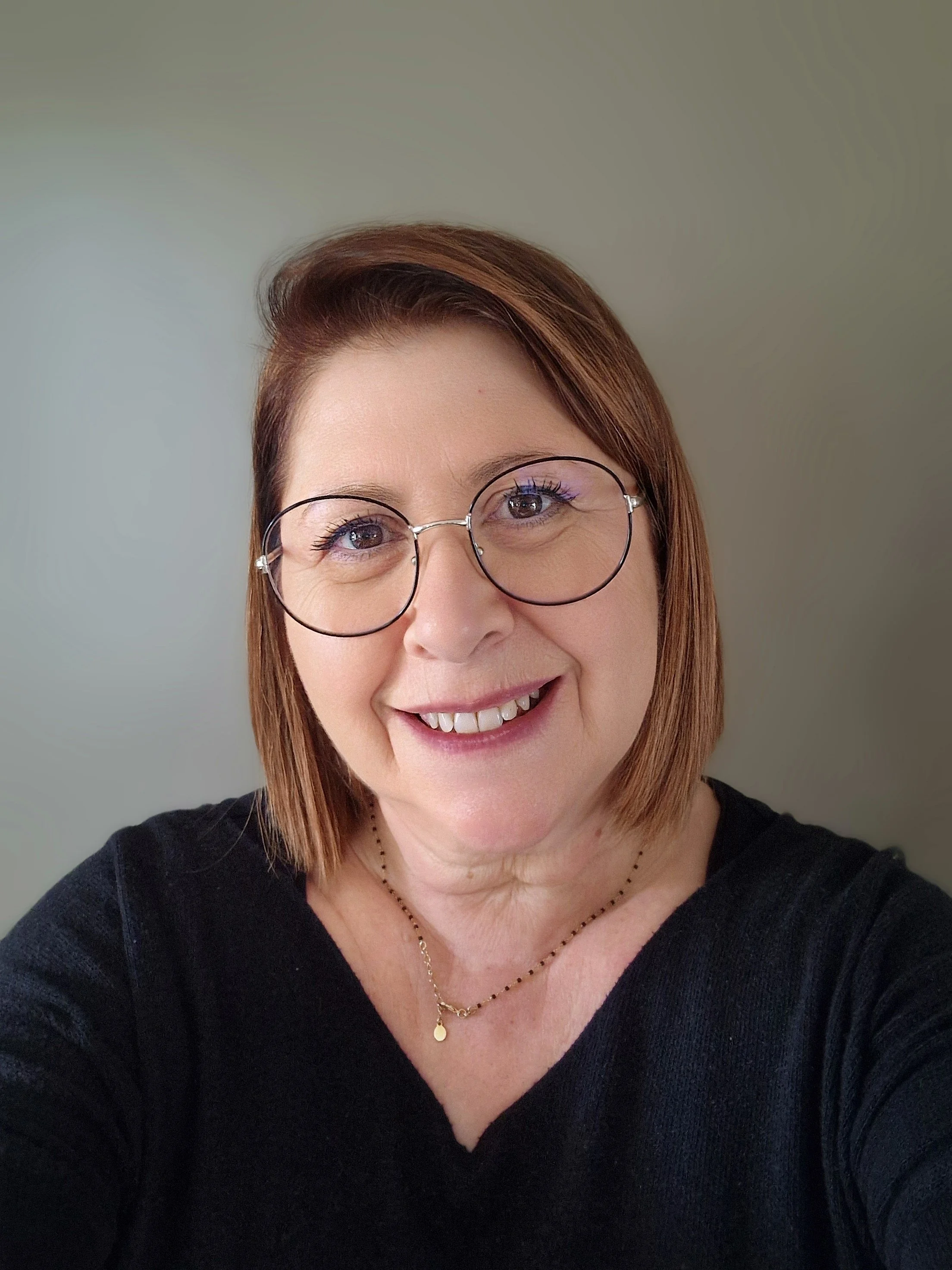 Portrait d'une femme souriante portant des lunettes rondes, un collier doré et un vêtement noir, avec un fond neutre.