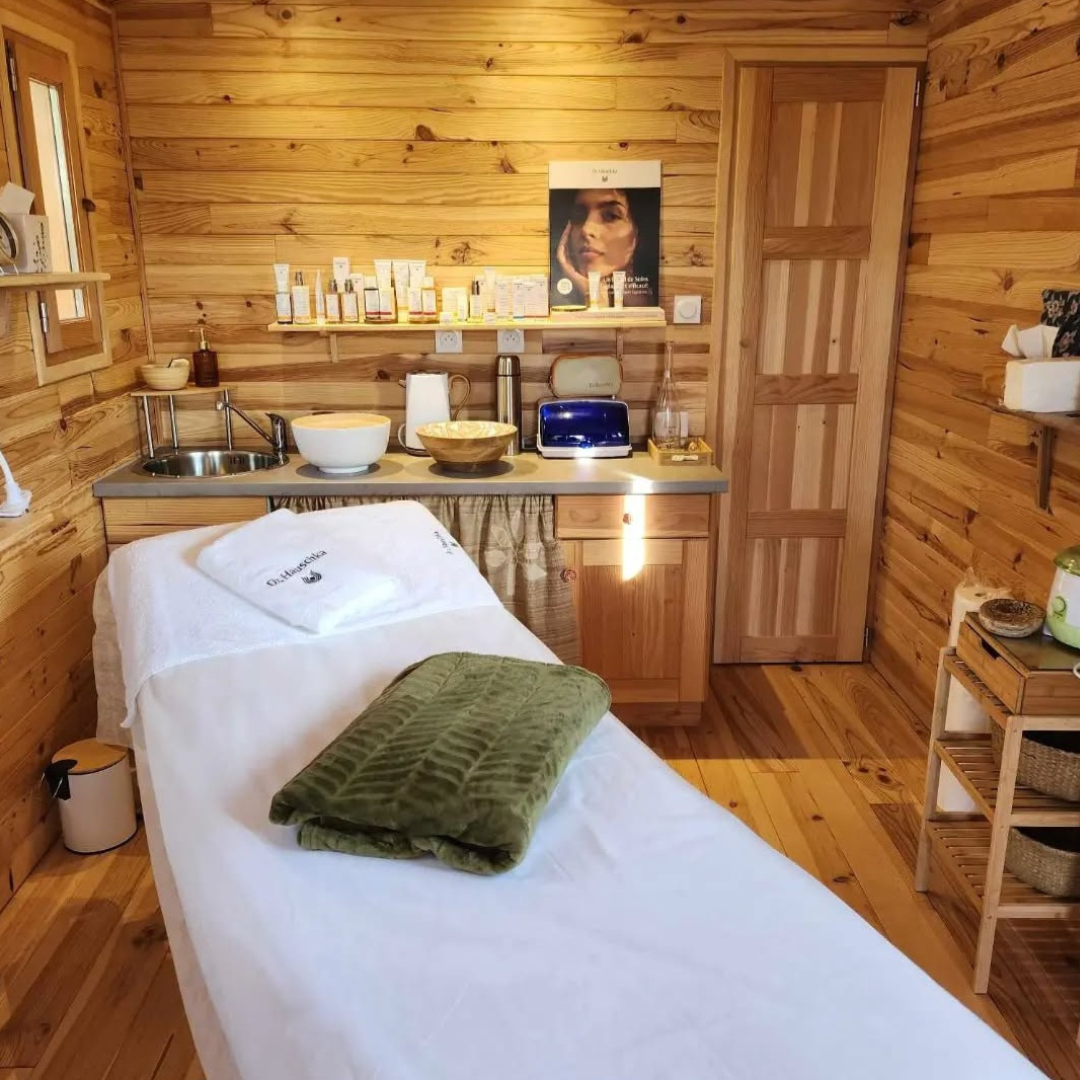 Cabine de massage avec murs en bois, lit de massage avec linge blanc et une serviette verte pliée, et une petite kitchenette avec un évier en acier inoxydable, diverses bouteilles de produits de soin, un grille-pain, et des accessoires ménagers.