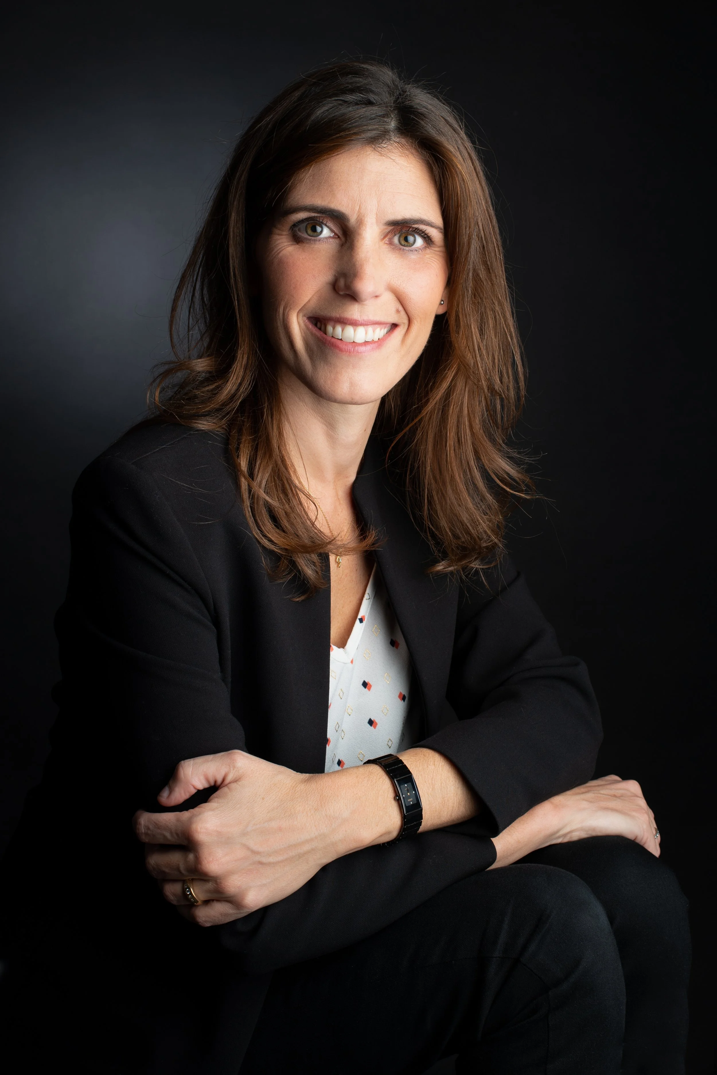Une femme souriante avec des cheveux bruns, portant un blazer noir et une montre, posant contre un fond noir.