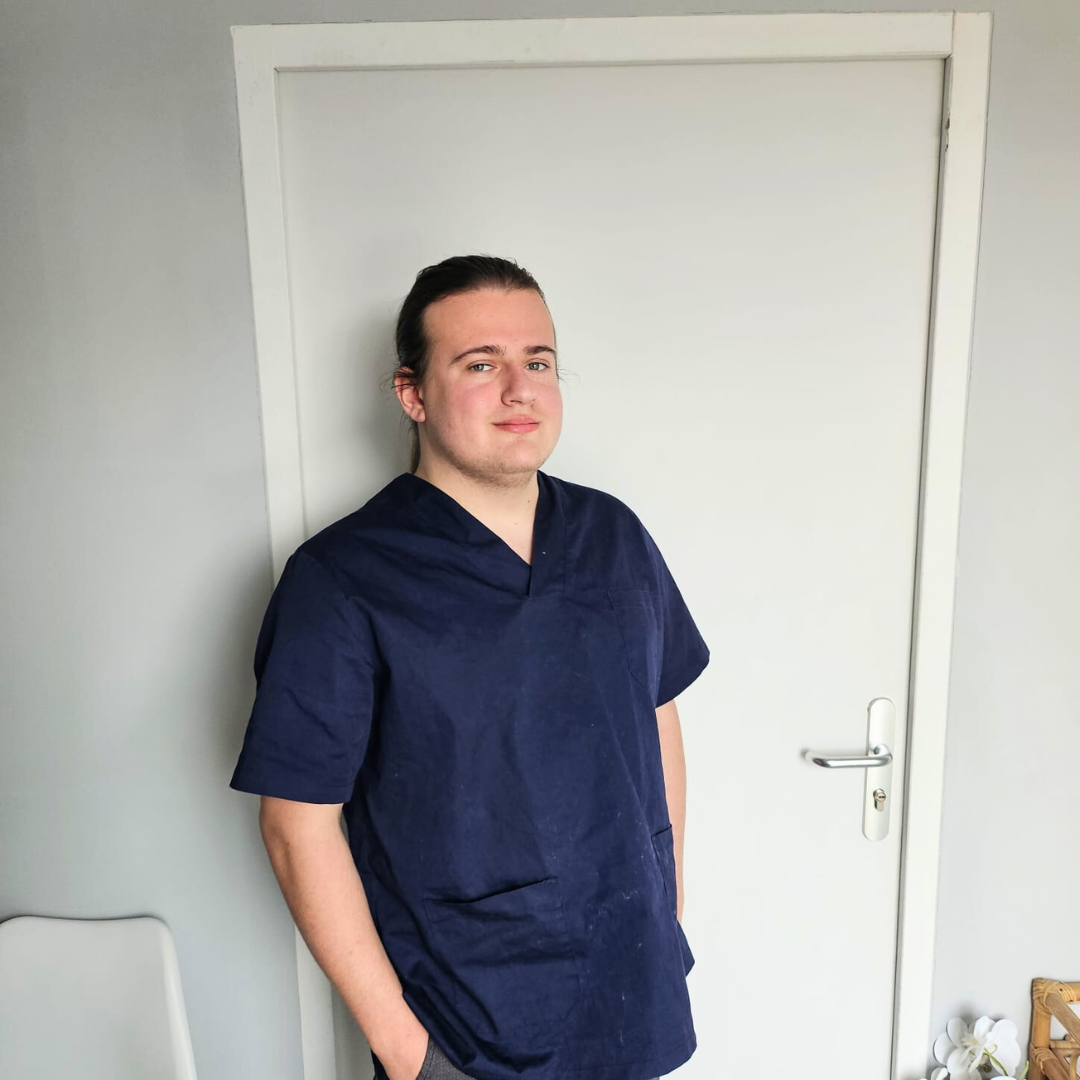 Jeune homme portant une blouse médicale bleue, debout devant une porte blanche, avec une expression neutre.