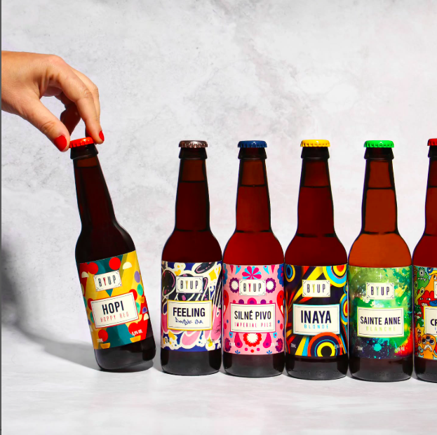 Une main attrape une bouteille de bière aux étiquettes colorées, alignées avec d'autres bouteilles de bière, sur une surface claire.