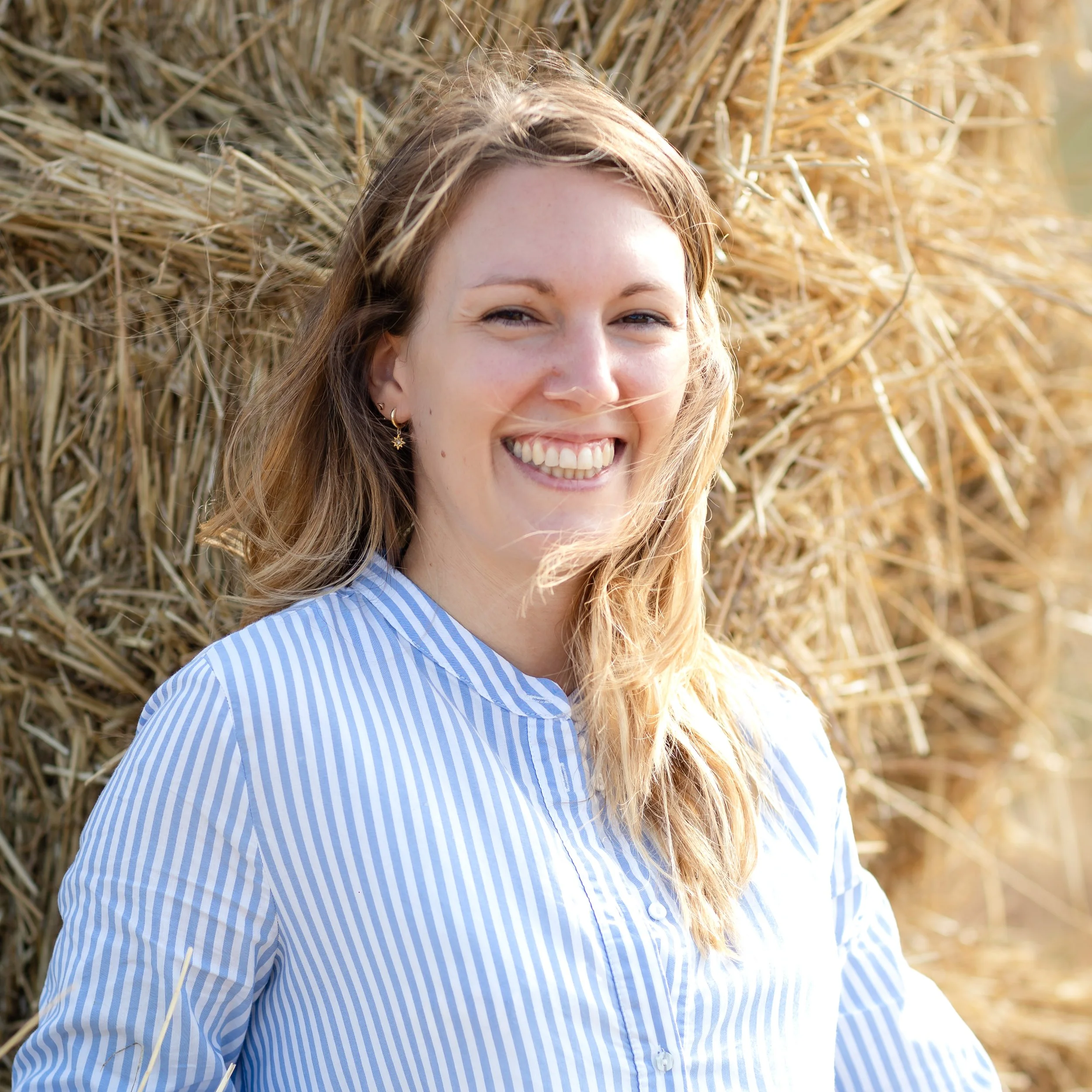 Jeune femme souriante avec des cheveux longs, portant une chemise bleue et blanche, posant près d'une pile de foin.