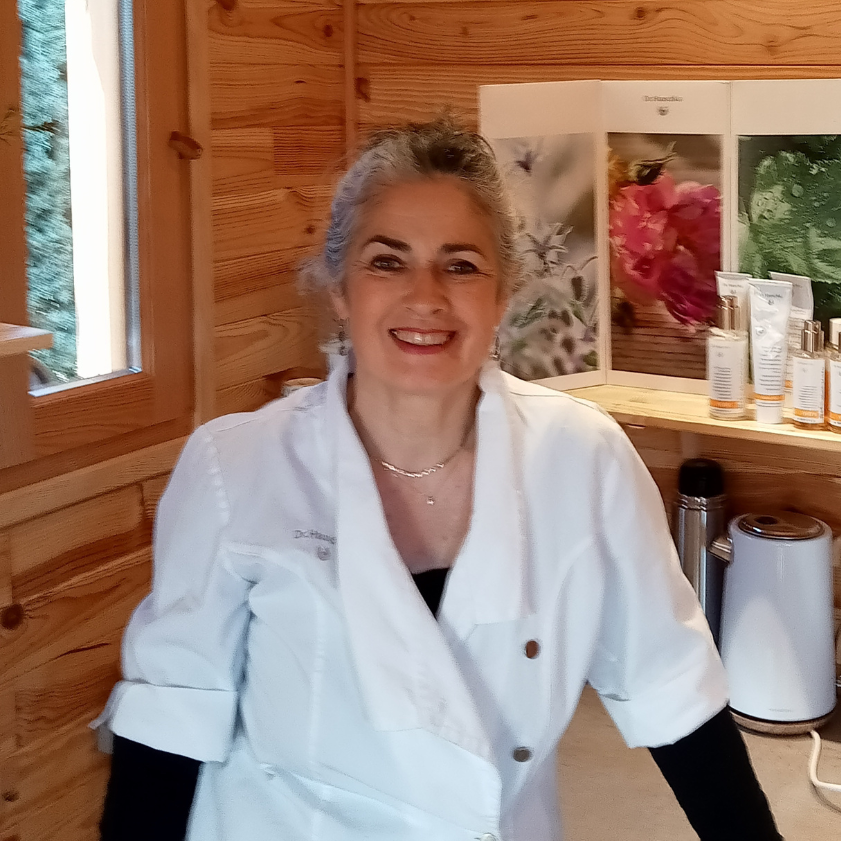 Une femme souriante portant un uniforme blanc dans une cuisine en bois, avec des produits de soins ou cosmétiques sur une étagère derrière elle.