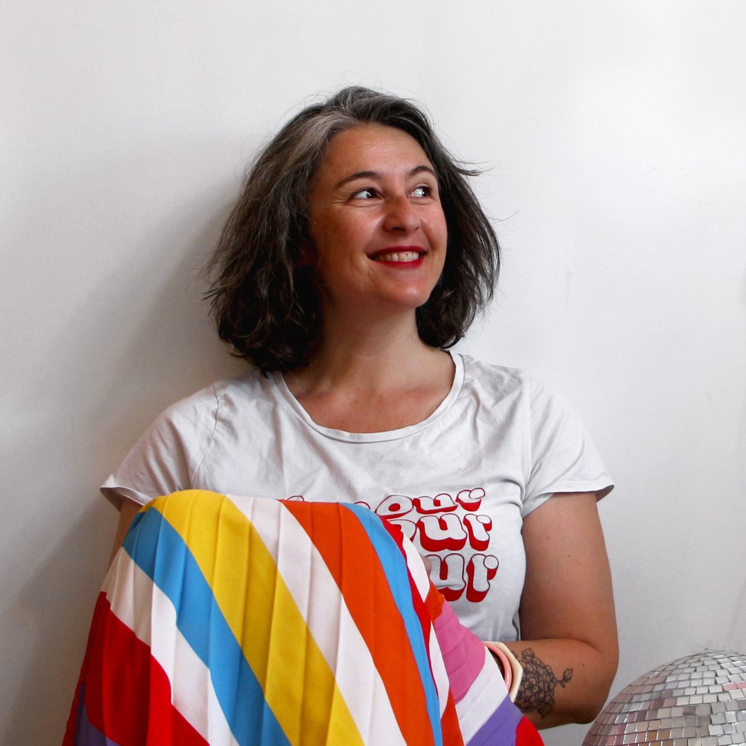 Une jeune femme souriante aux cheveux bouclés, portant un t-shirt blanc avec un design rouge, tient un coussin coloré avec des rayures diagonales dans des couleurs vives.