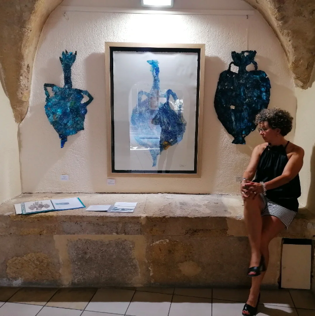 Une femme assise devant une œuvre d'art représentant la silhouette d'une femme, accompagnée de deux autres Artworks en forme de silhouettes féminines en mosaïque, exposée sur un mur dans une galerie.