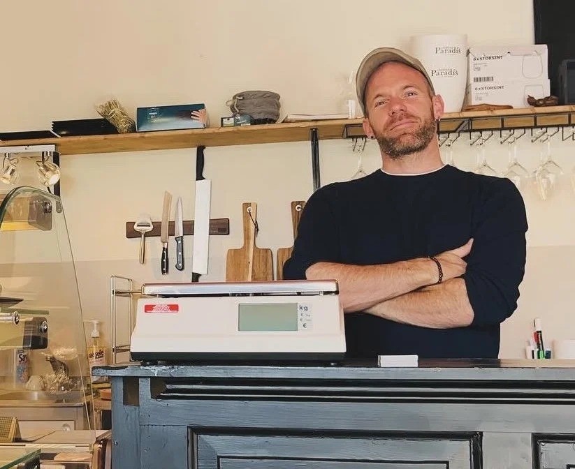 Homme debout derrière un comptoir de café ou boutique, avec une balance électronique, bras croisés, se trouvant dans une cuisine ou un espace de vente avec des ustensiles de cuisine accrochés en arrière-plan.