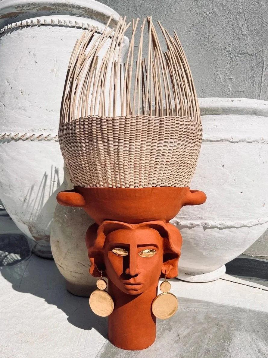 Une sculpture en terre cuite d'une tête de femme portant des boucles d'oreilles, dont le sommet est magnifiquement orné de matériaux tressés. La sculpture est placée contre un mur blanc avec plusieurs poteries en arrière-plan.