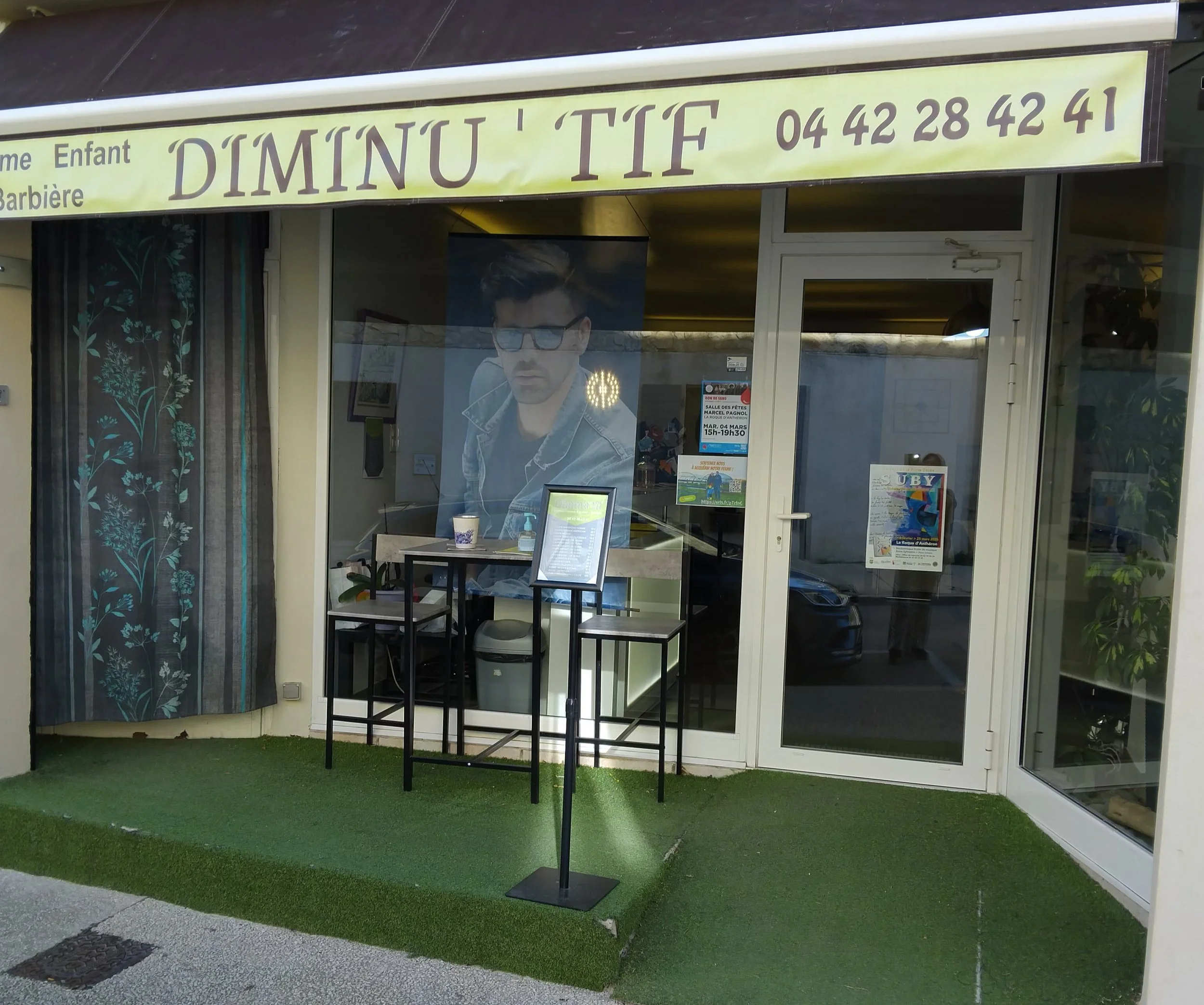 Facade d'un salon de coiffure appelé 'DIMINU'TIF' avec un grand portrait d'un homme portant des lunettes de soleil à l'intérieur, une enseigne jaune avec le nom et le numéro de téléphone en haut, et des petites tables avec des menus en dehors, sur une surface en faux gazon.