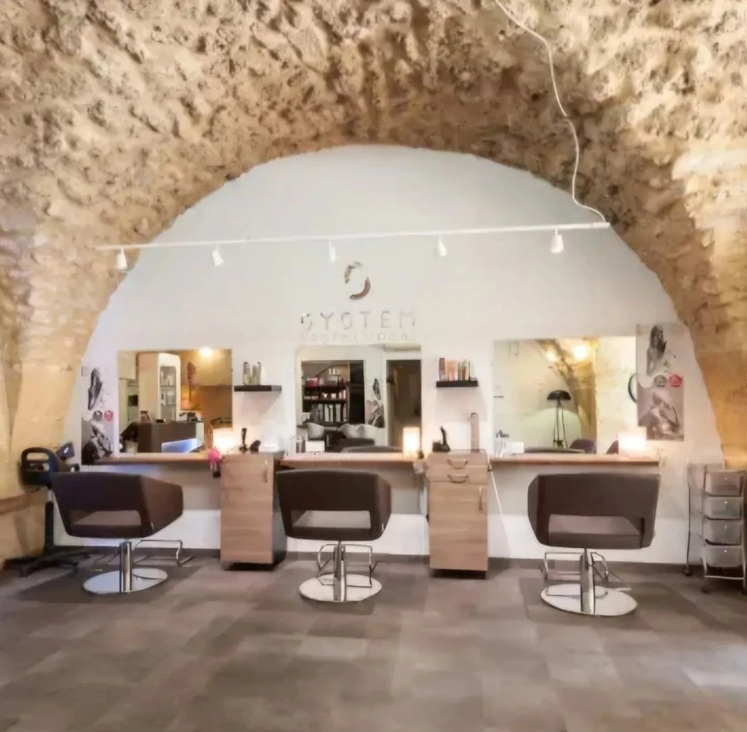 Salon de coiffure avec trois fauteuils devant un miroir au mur blanc et un plafond en pierre.