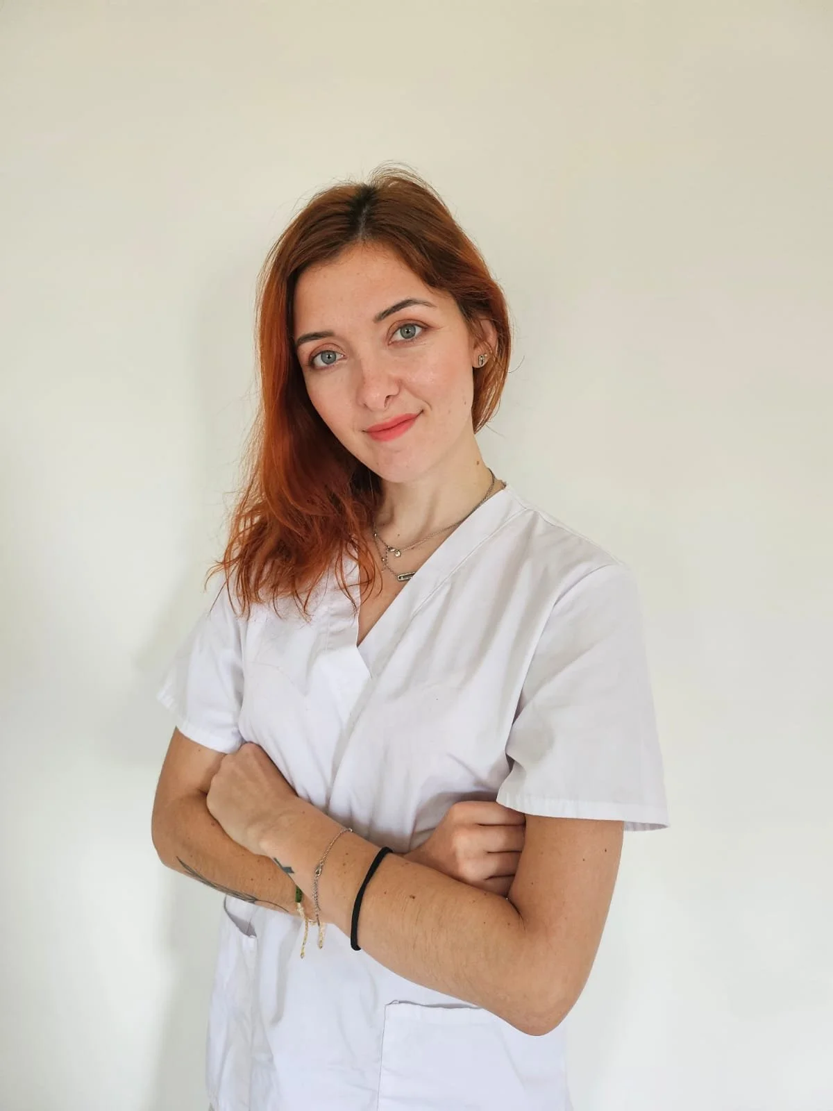 Jeune femme à cheveux roux et yeux bleus portant une blouse blanche, souriant et posant avec les bras croisés contre un mur blanc.