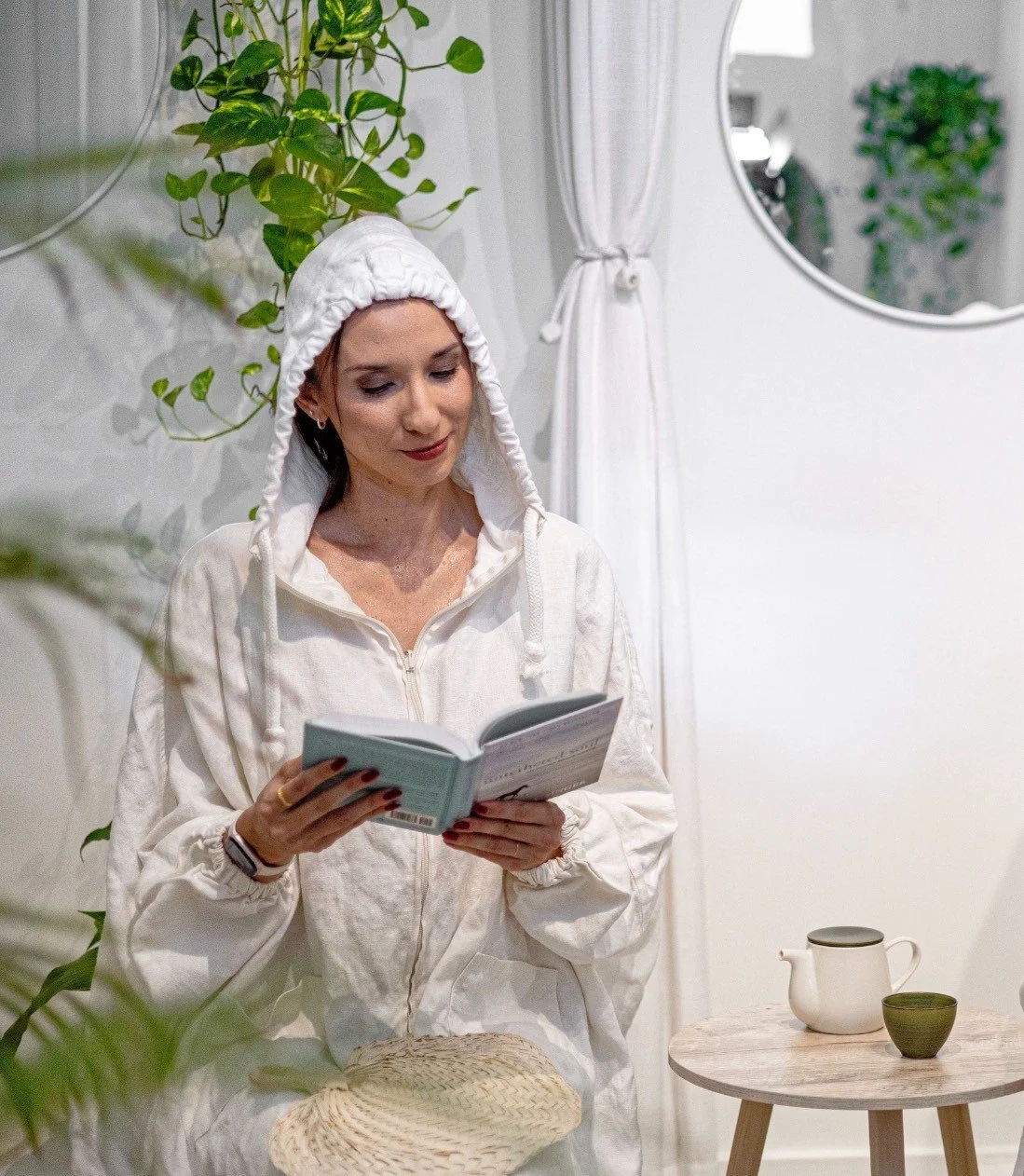 A woman wearing a white hoodie with the hood up, reading a book indoors. There is a small round wooden table with a teapot and a cup next to her, and greenery decor surrounding her.
