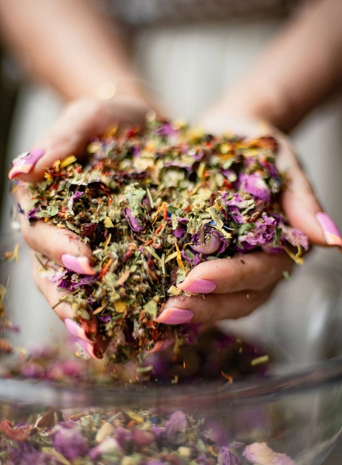 Hands holding a handful of dried herbal tea leaves with purple, yellow, and green plant fragments, ready to steep.