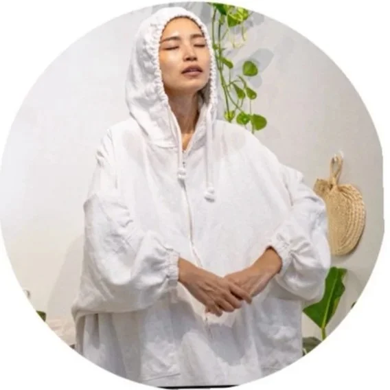 A woman in a white hooded spa or loungewear tunic with her hands clasped in front of her, standing in a room with plants and decorative items.