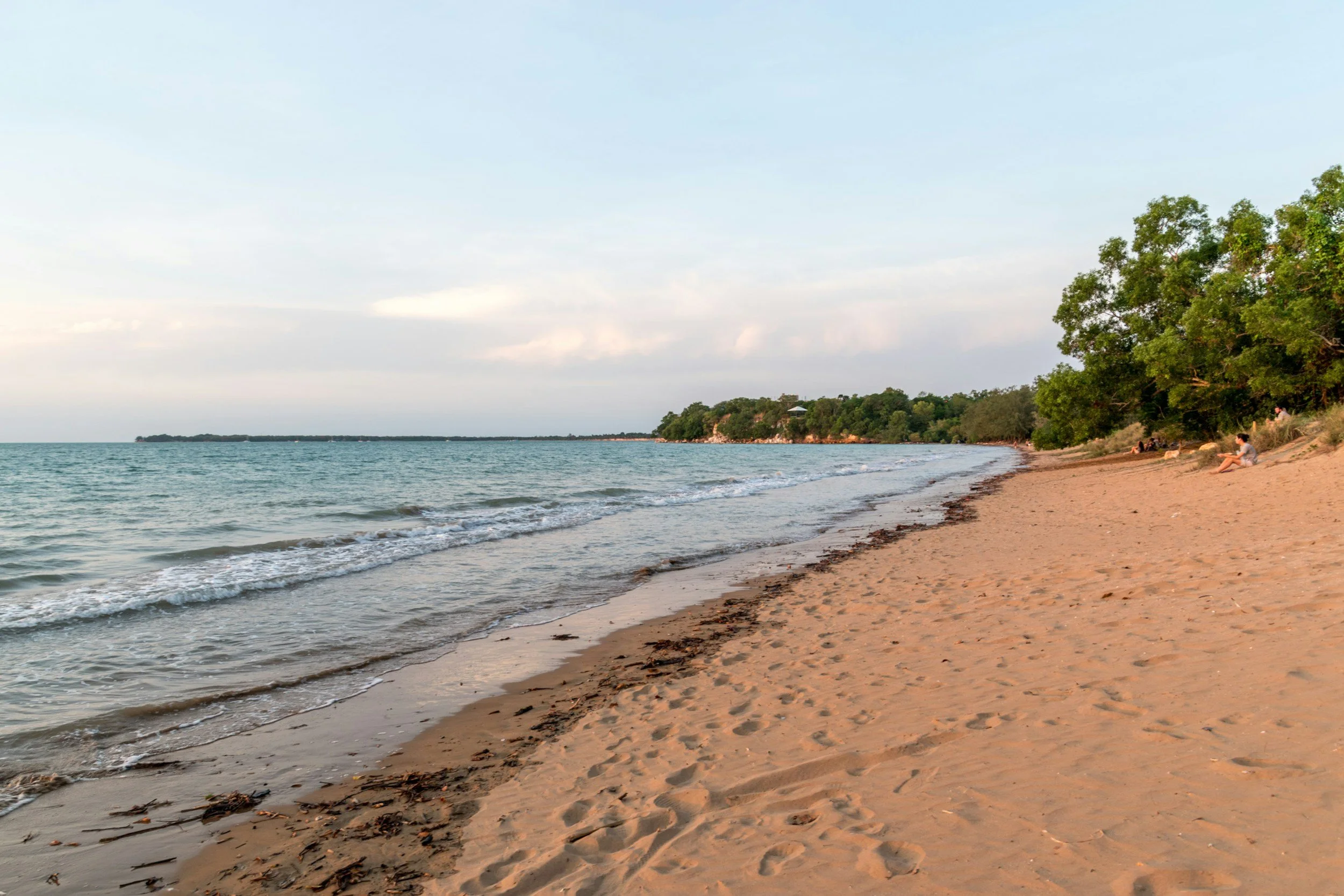 A sandy beach with a few people sitting near the water, green trees in the background, and the sky is partly cloudy.
