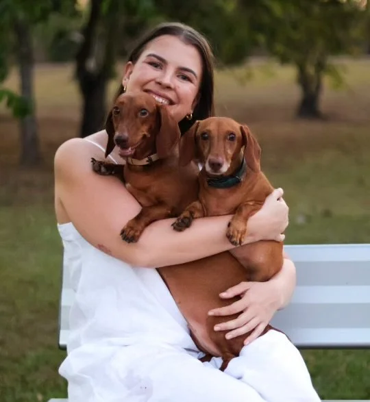 A young woman smiling and hugging two small brown dachshund dogs outdoors in a park.
