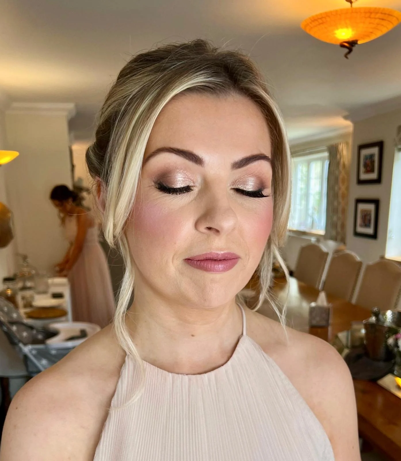 A woman with styled blonde hair and makeup, wearing a beige dress with thin straps, standing in a warmly lit room with framed pictures and a window in the background.