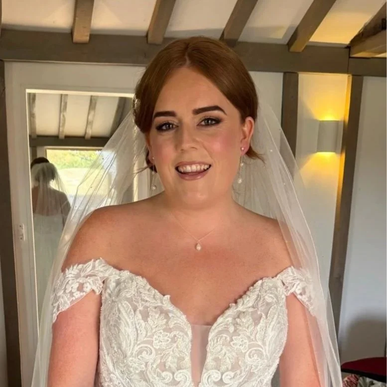 A bride with reddish-brown hair styled in an updo, wearing a white lace off-the-shoulder wedding gown and a sheer veil. She has earrings, a necklace, and is smiling at the camera, standing inside a room with wooden beams and warm lighting.