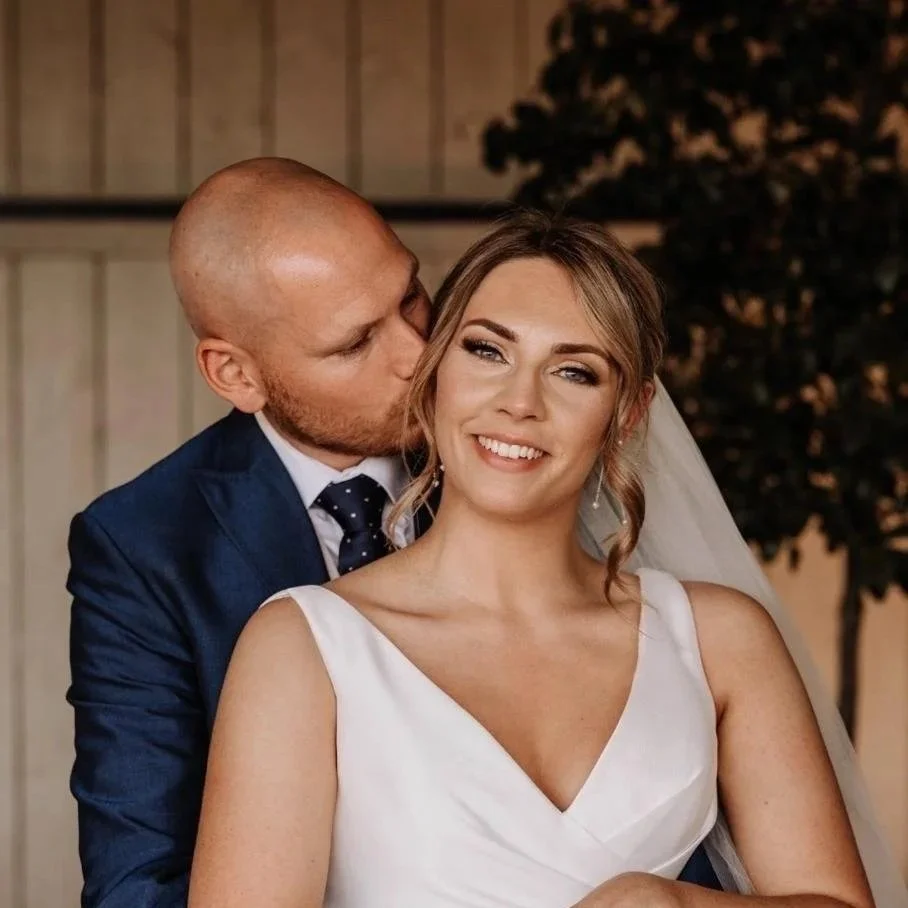 A groom kissing his bride on the forehead at their wedding, both smiling warmly, with the bride wearing a white dress and veil, inside a decorated indoor venue.