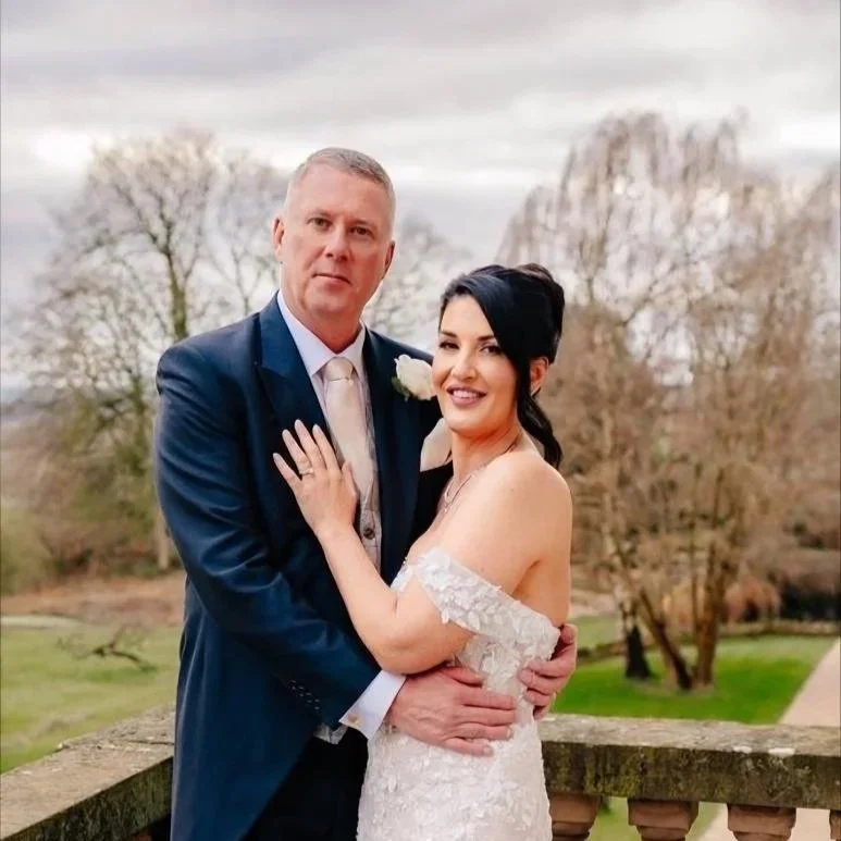 A couple dressed in wedding attire, standing outdoors on a balcony with trees in the background. The man wears a blue suit and white shirt, and the woman wears a white off-shoulder wedding gown. The woman is smiling, and the man has a neutral express