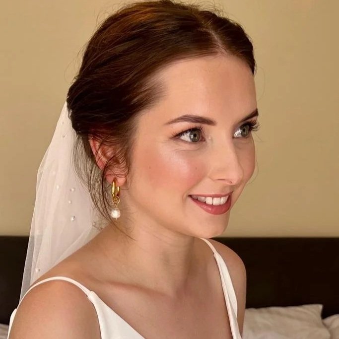 A smiling woman with brown hair, wearing a wedding veil and gold earrings with pearls, dressed in a white wedding gown.