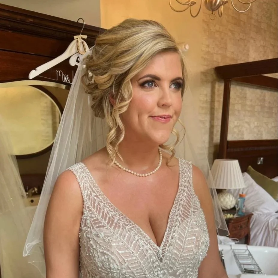A bride with blonde hair styled in soft curls, wearing pearl earrings and a pearl necklace, dressed in a lace wedding gown with beaded embroidery, standing in a bedroom with a wooden headboard, a mirror, and a bed with white linens.