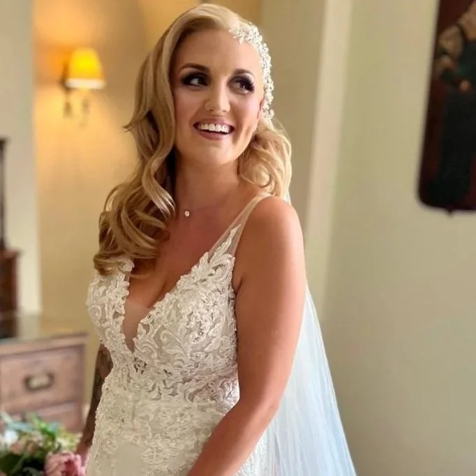 A bride in a lace wedding dress with a low neckline, smiling in a softly lit room.