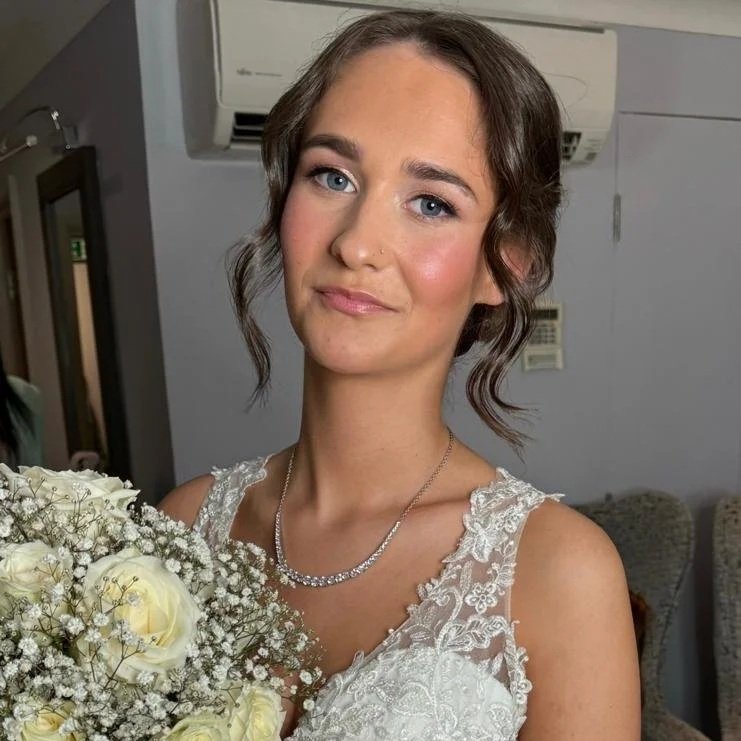 A young woman in a white lace wedding dress holding a bouquet of white roses and baby's breath, smiling softly indoor setting.
