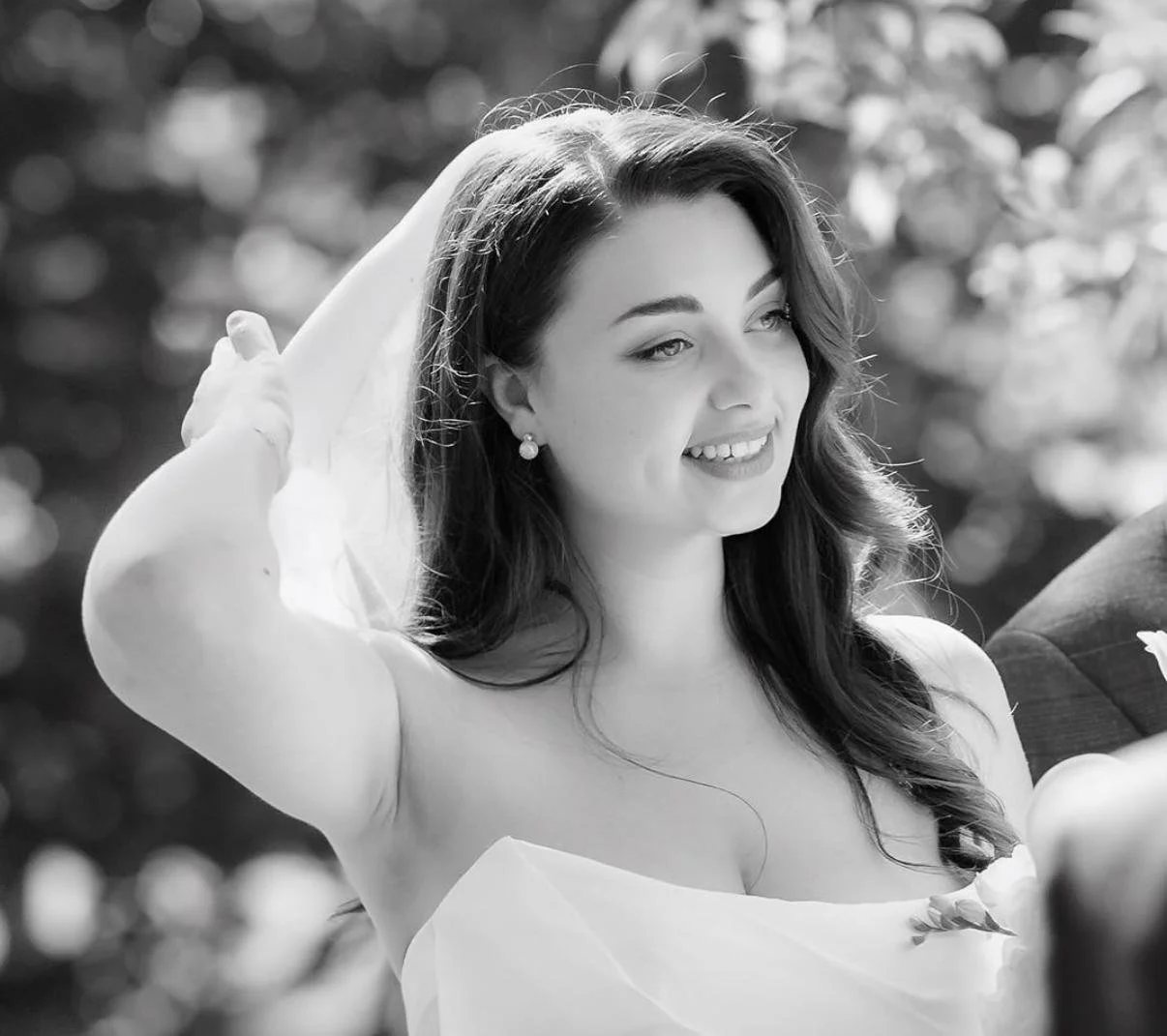 A black and white photo of a woman with long dark hair wearing pearl earrings, smiling, with her hand behind her head, outdoors with blurred trees in the background.