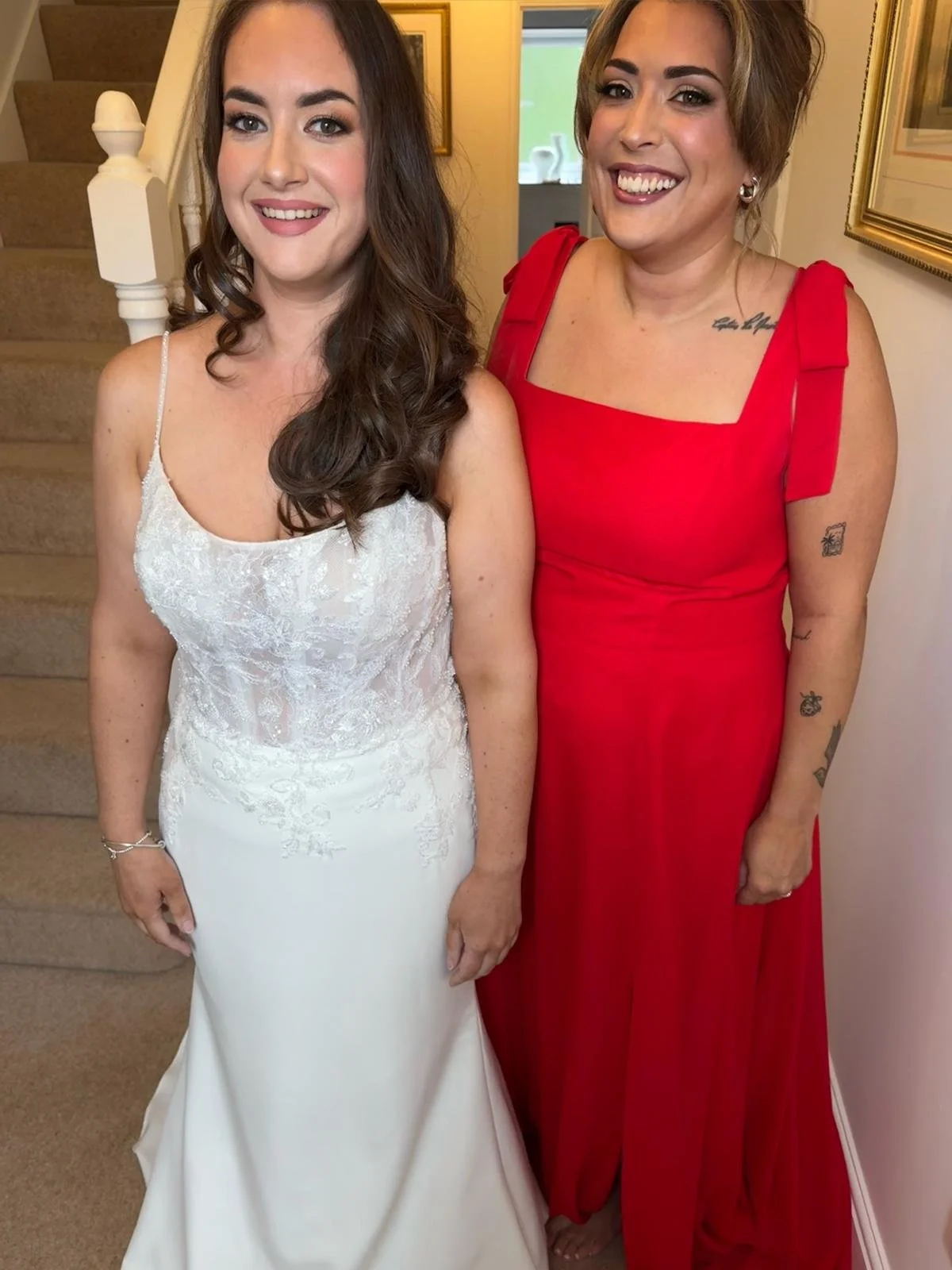Two women standing together smiling, one in a white wedding dress and the other in a red dress, inside a house near a staircase.