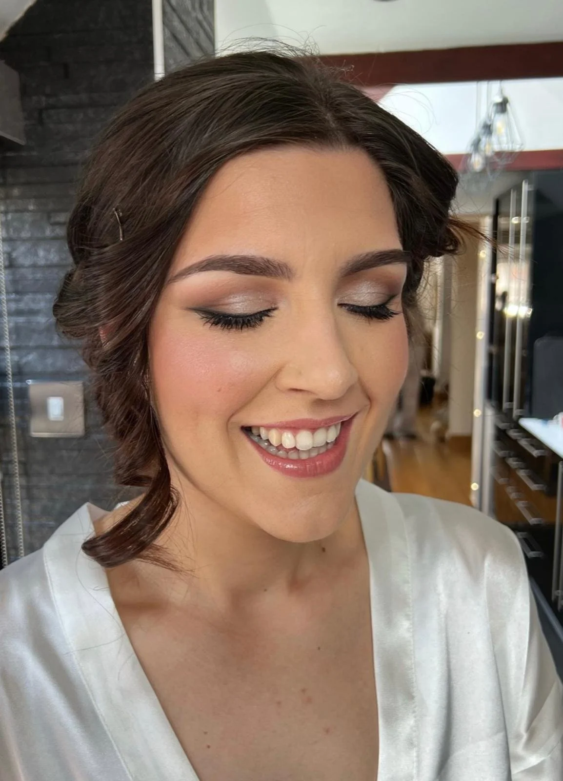 A woman with short brown hair, makeup, and a white satin top smiling with closed eyes, indoors with a modern decor background.