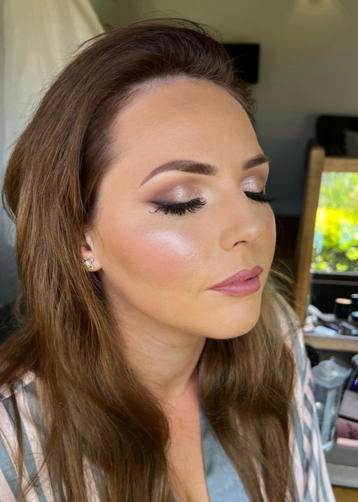 Close-up of a woman with makeup, closed eyes showing shimmery eyeshadow and long lashes, with light pink lipstick, and wearing earrings, in a room with a window and makeup tools in the background.