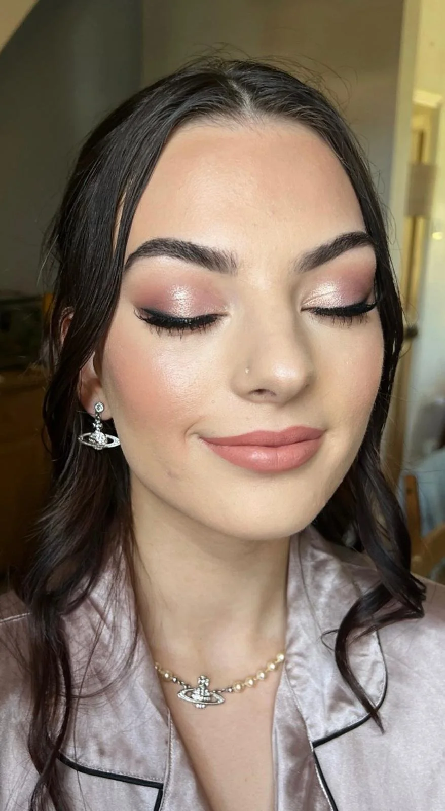 A young woman with closed eyes, wearing makeup including shimmery eyeshadow, eyeliner, and peach lipstick, plus earrings and a necklace, smiling gently.