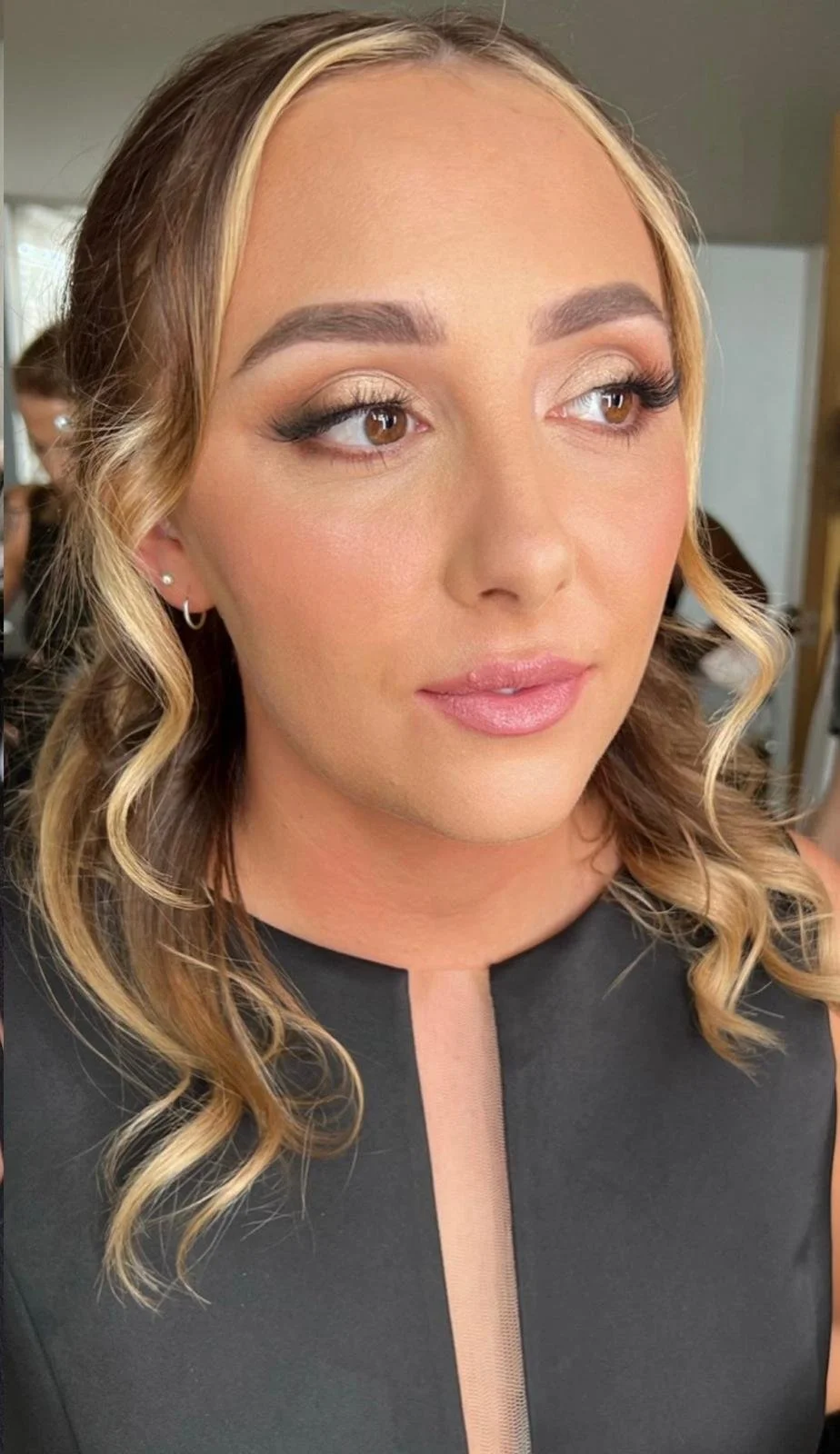 Close-up of a woman with wavy blonde hair, light makeup, and a black sleeveless top with a beige sheer panel in the front.