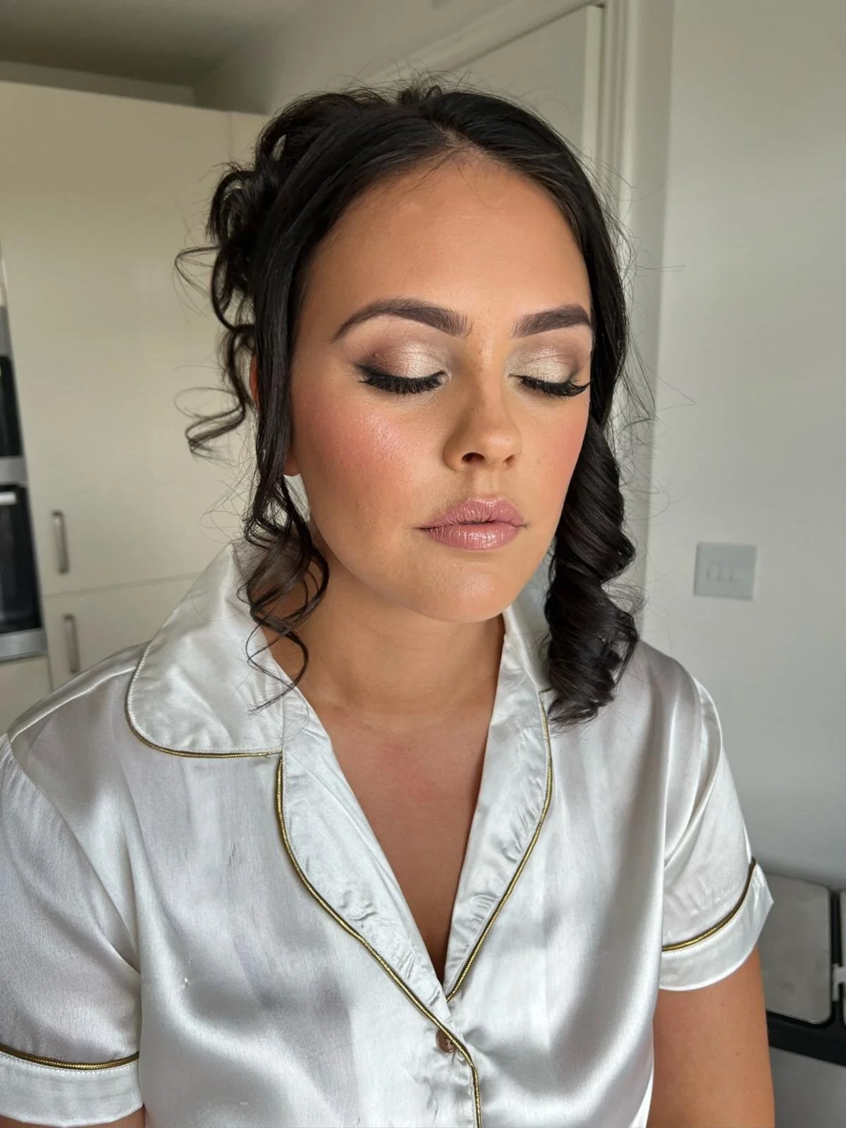 A woman with dark, curly hair styled in loose waves, wearing gold and beige eyeshadow, eyeliner, mascara, and neutral-colored lipstick, dressed in white satin pajamas with gold piping, standing in a modern kitchen with a white wall and appliances in 