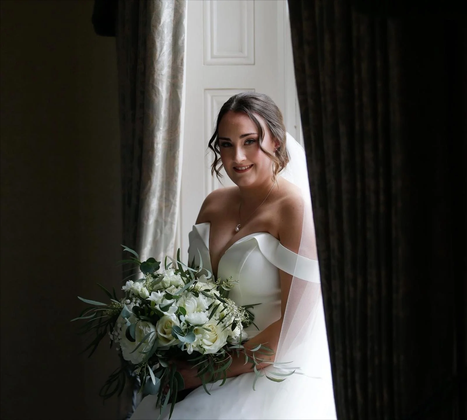 A lovely smiling bride of Kelly Marie Makeup, holding a bouquet of white flowers.
