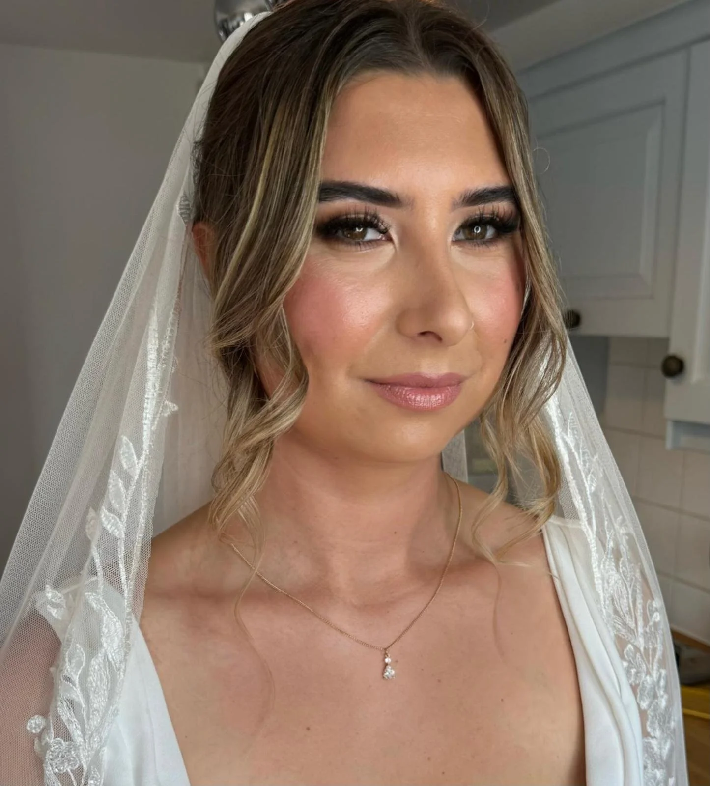 Close-up of a woman with fair skin, wearing makeup with highlighting eyes and glossy lips, dressed in a wedding gown with a lace veil and a delicate necklace, standing in a room with white cabinetry in the background.