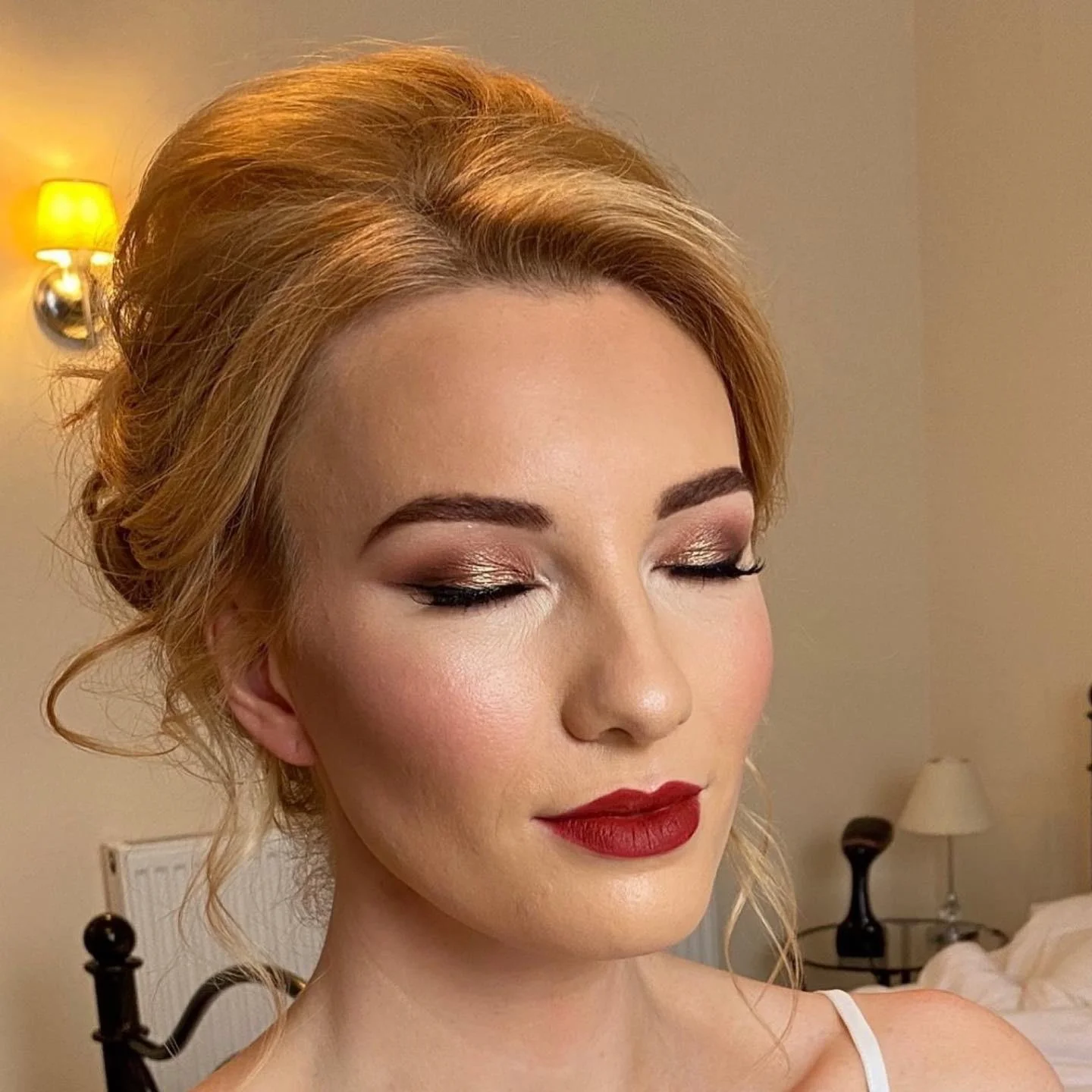 A woman with styled blonde hair and makeup including gold eyeshadow and red lipstick, in a bedroom with beige walls, a black lamp on a nightstand, and a bed with white bedding.