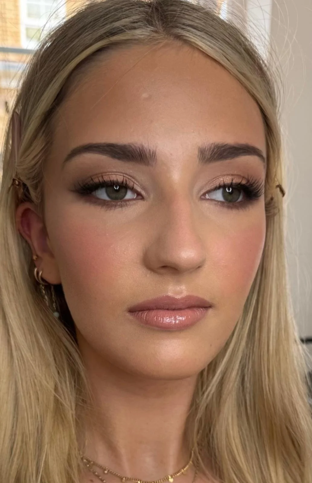 Close-up of a woman with blonde hair, green eyes, and makeup with neutral tones, wearing multiple earrings and a gold necklace.