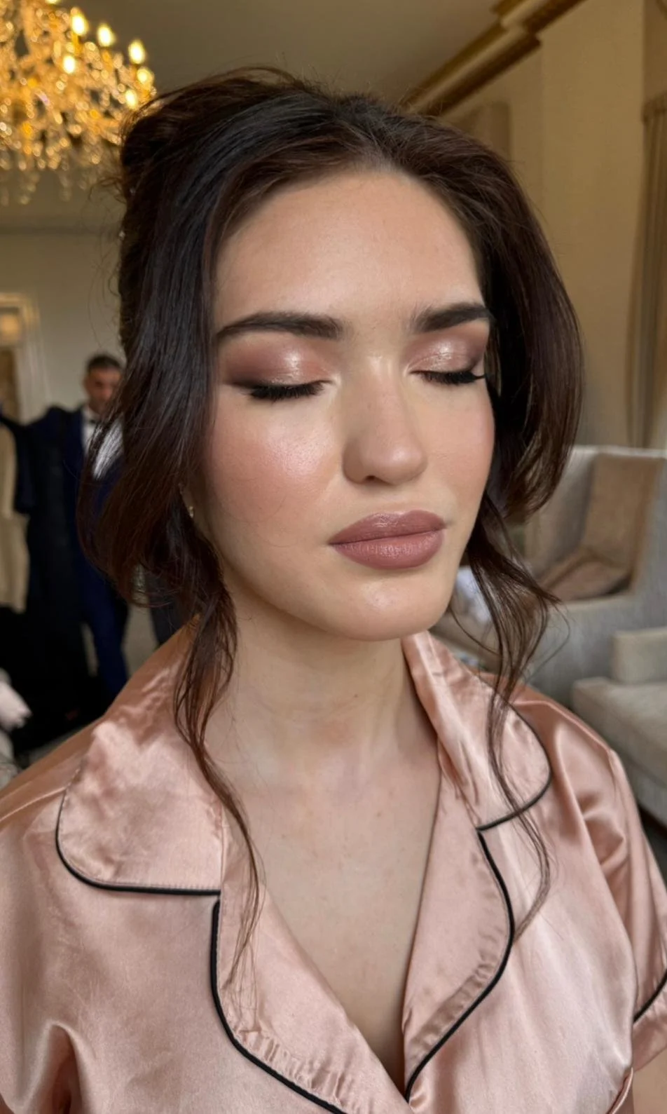A woman with closed eyes and styled dark hair, wearing makeup and a satin pink garment, standing in an elegant room with a chandelier in the background.