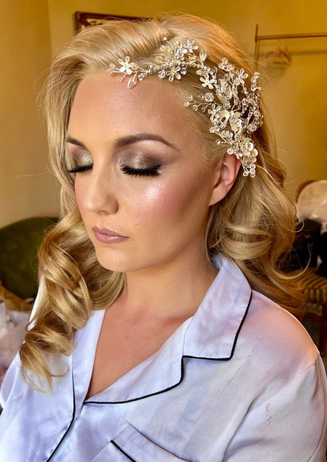 A woman with blonde hair styled in loose curls wearing a silver floral headpiece, with silver eyeshadow, black eyeliner, long eyelashes, and glossy lips, dressed in satin pajamas with black piping.