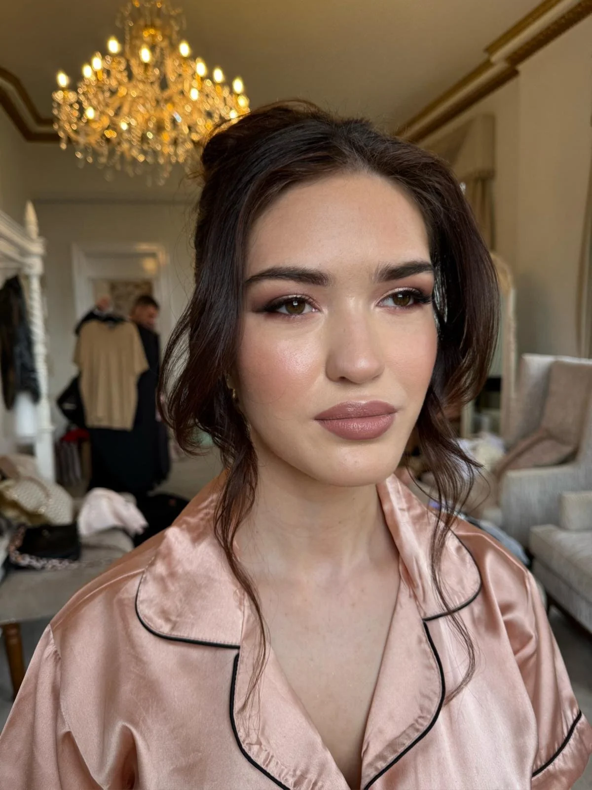 A woman with dark wavy hair styled in loose waves, wearing makeup, a satin pink robe with black piping, is in a luxurious room with a chandelier, a mirror, and furniture. She has a neutral expression and looks slightly to the side.