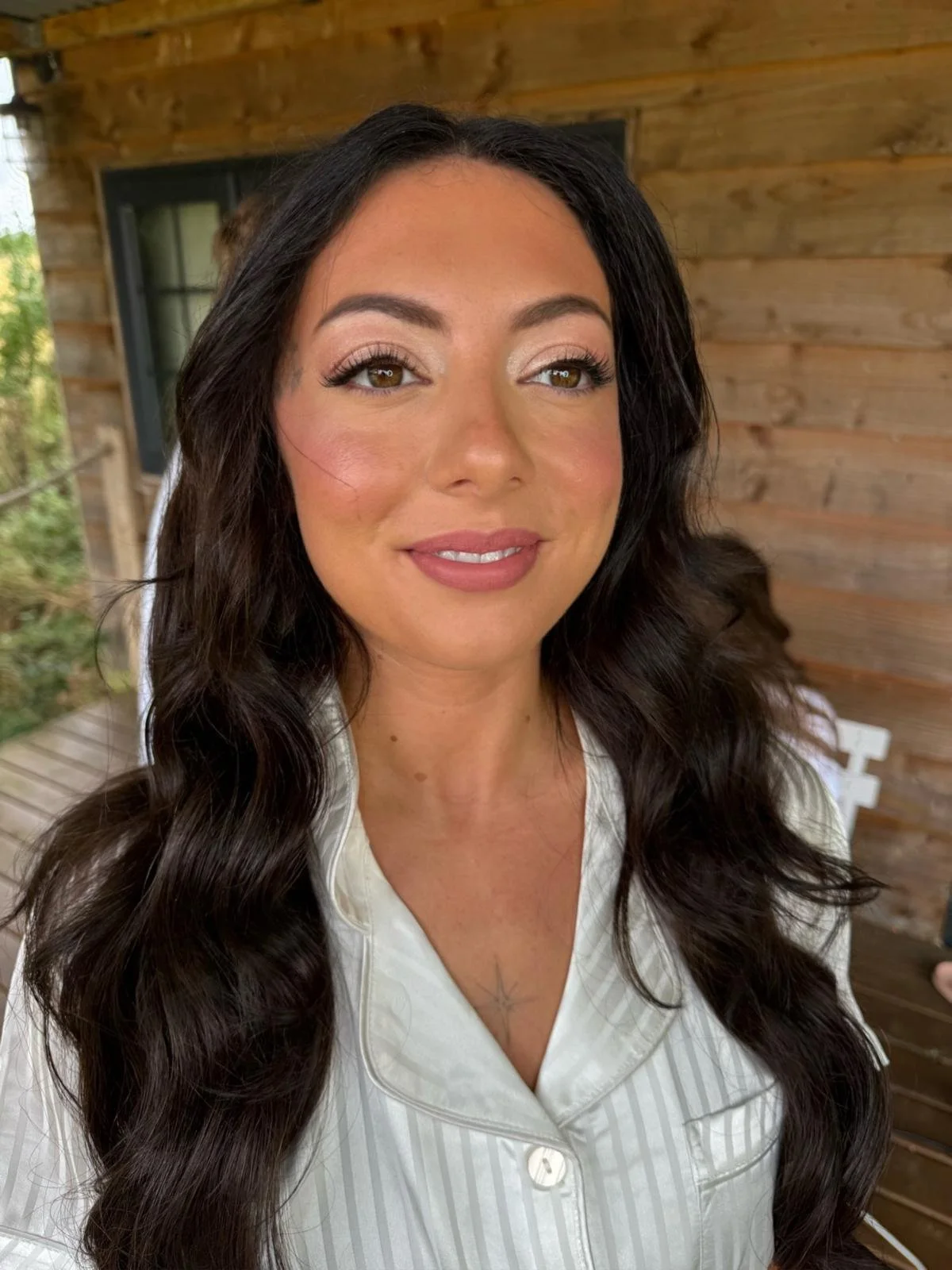 A woman with long dark wavy hair smiling, wearing light-colored striped pajamas, standing in front of a wooden wall with trees visible outside a window.